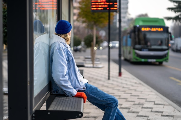 Zdjęcie tysiącletni facet siedzący na ławce na przystanku autobusowym i patrzący na przyjeżdżający autobus