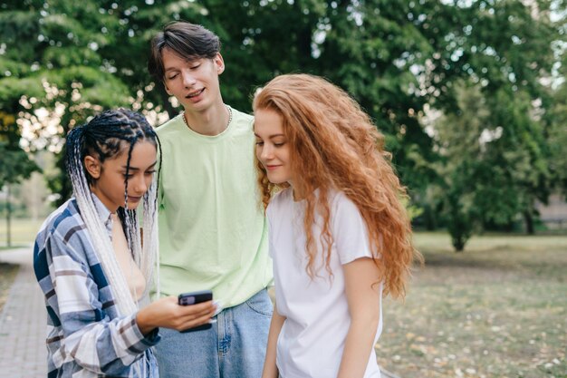 Tysiącletnia grupa blogerów stojących w parku i oglądających trendy wideo na nowoczesnym gadżecie telefonu komórkowego Styl życia młodzieży Media społecznościowe Blog wideo na żywo na ekranie telefonu komórkowego