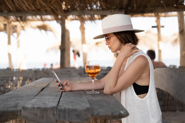 Tysiącletnia kobieta siedzi w tropikalnym barze przy drewnianym stole w okularach przeciwsłonecznych, a kapelusz trzyma w dłoniach smartfon, przeglądając kanał informacyjny Wakacje w krajach tropikalnych