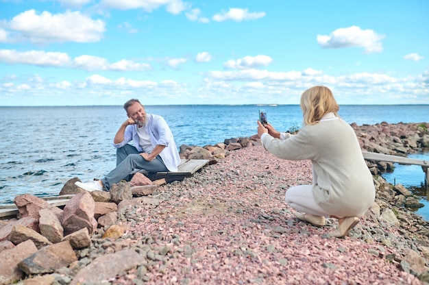 Uchwyć chwilę. Blond kobieta kucająca ze smartfonem fotografująca brodatego mężczyznę siedzącego na ziemi w pobliżu morza