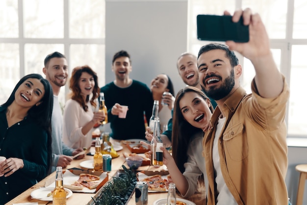 Uchwyć chwilę. Grupa młodych ludzi w strojach casualowych robi selfie i uśmiecha się podczas kolacji w pomieszczeniu