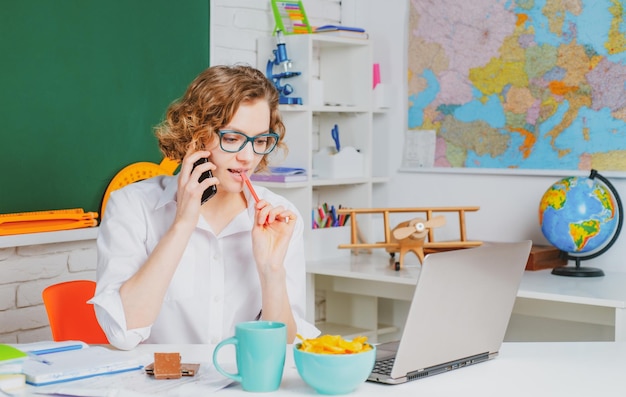 Ucz się z zabawną zabawną nauczycielką z telefonem komórkowym, używając laptopa siedzącego w swoim miejscu pracy i nauczającego