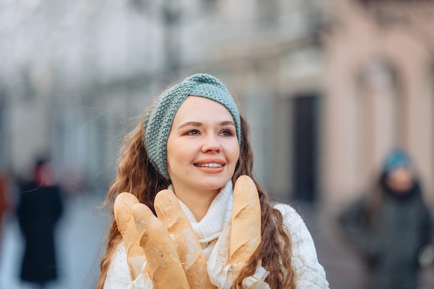 Uczciwa stylowa dziewczyna pozuje do reklamy produktów piekarniczych w chłodnej odzieży, trzymając w rękach 4 opakowania bochenków. Spojrzenie w bok i miły uśmiech. Rozmyte tło centrum miasta.