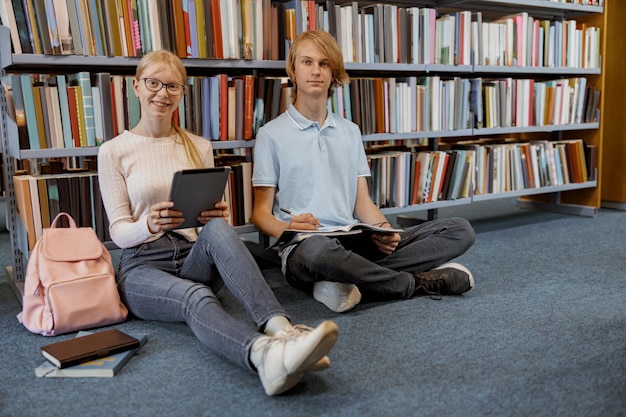 Uczeń przyjaciół siedzi na podłodze w pobliżu półek z książkami w bibliotece i studiuje, przygotowując się do egzaminu