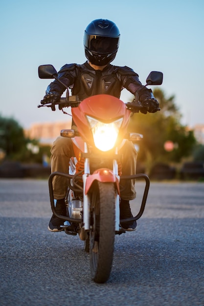Uczeń w kasku pozuje na motocyklu, widok z przodu, wieczorna lekcja w szkole motocyklowej. Szkolenie początkujących motocyklistów, rowerzystów ćwiczących na motordromie w szkole motorowej