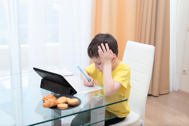 Uczeń w żółtej koszulce siedzi przy stole z cyfrowym tabletem studiującym w domu