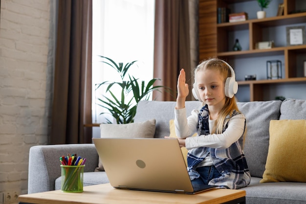 Zdjęcie uczennica podstawowa nosząca słuchawki prowadzi rozmowę wideo online z nauczycielem podnoszącym rękę, aby odpowiedzieć na pytanie urocze dziecko uczące się w domu za pomocą laptopa nauka na odległość edukacja domowa