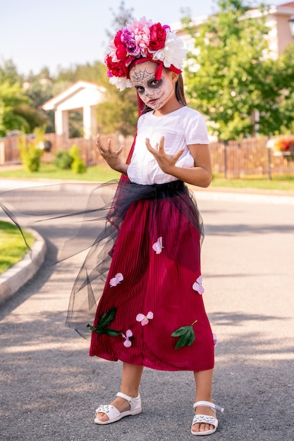 Uczennica z pomalowaną twarzą w kostiumie na halloween, trzymająca się za ręce, patrząc na ciebie z nadąsaną miną w słoneczny dzień