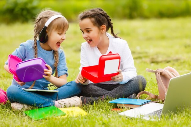 Uczniowie szkoły podstawowej z pudełkami na lunch w rękach. Dziewczyny z plecakami jedzą na trawniku. Początek lekcji. Pierwszy dzień jesieni.