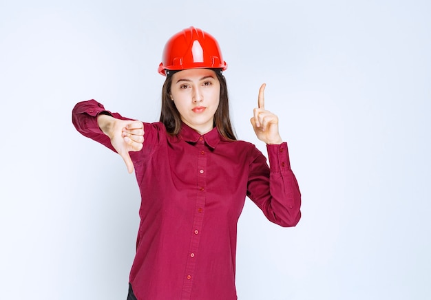 Udany architekt żeński w czerwonym twardym kasku stoi i daje kciuki w górę w dół.