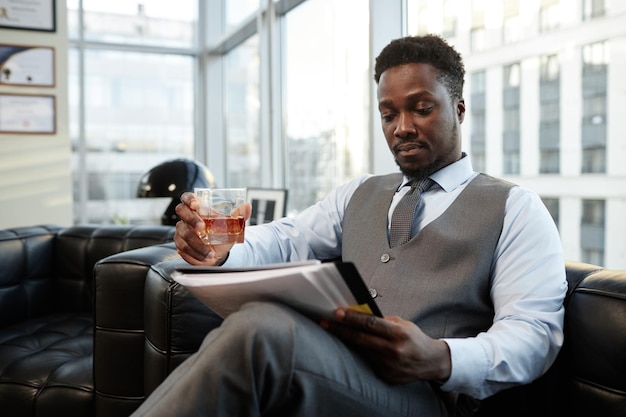 Udany czarny biznesmen pije whisky w biurze i czyta dokumenty.