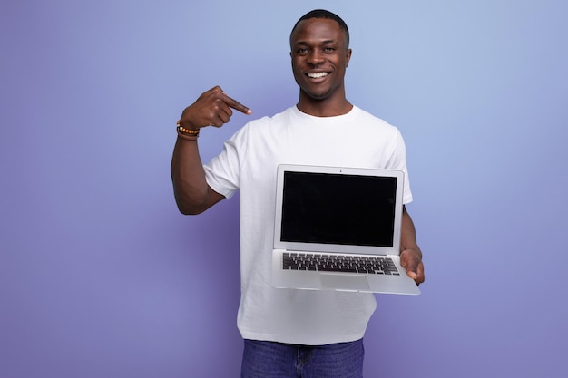 Udany młody afrykański mężczyzna w białej koszulce pracujący zdalnie za pomocą laptopa na tle studia z