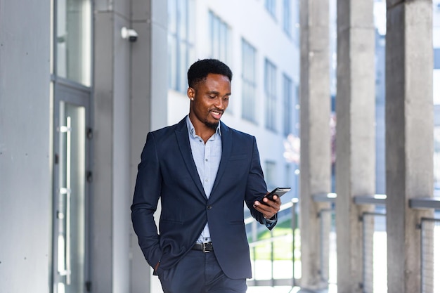 Udany młody pewny siebie African American biznesmen poruszający się z telefonem komórkowym w rękach na tle nowoczesnego biurowca w centrum miasta czarny biznesmen spacery za pomocą telefonu komórkowego w formalnym garniturze