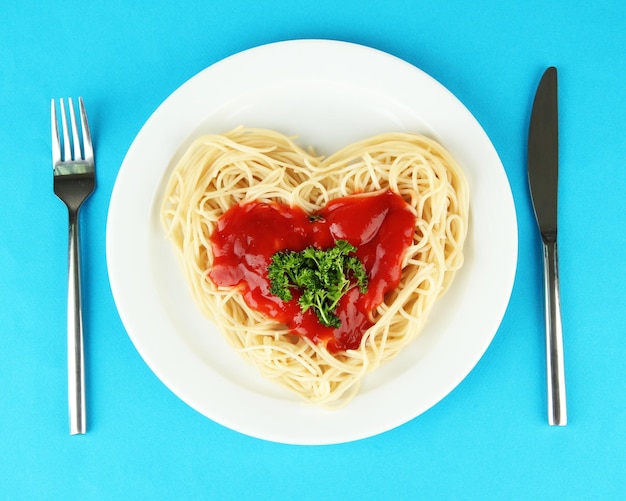 Ugotowane spaghetti starannie ułożone w kształcie serca i polane sosem pomidorowym, na kolorowym tle