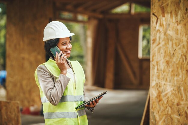 Ujęcie afrykańskiej architektki za pomocą smartfona i sprawdzanie placu budowy nowego drewnianego domu. Ubrana jest w ochronną odzież roboczą i biały kask.