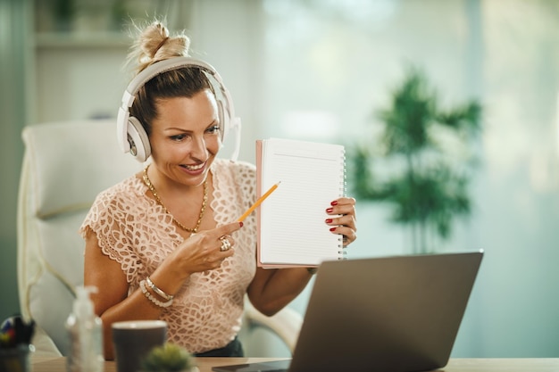 Ujęcie atrakcyjnej młodej bizneswoman siedzącej samotnie w swoim domowym biurze ze słuchawkami i pracującej na laptopie podczas pandemii COVID-19.