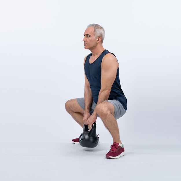 Ujęcie całej długości ciała, atletyczny i sportowy starszy mężczyzna robi przysiad z kettlebell do treningu ciała na odizolowanym tle Zdrowa, aktywna sylwetka i styl życia związany z pielęgnacją ciała po przejściu na emeryturę Clout