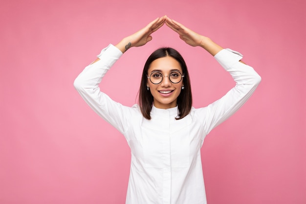 Ujęcie całkiem pozytywnej uśmiechniętej młodej brunetki ubranej w białą koszulę i stylowe okulary optyczne odizolowane na różowym tle, patrząc na kamerę i pokazując znak gestu domu