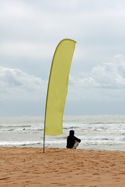 Ujęcie Człowieka Siedzącego Na Plaży Oglądania Fal Obok Dużej Flagi.