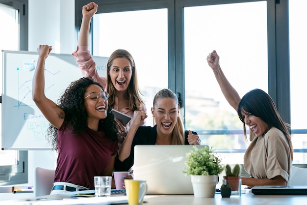 Ujęcie Czterech Szczęśliwych, Inteligentnych Kobiet Biznesu Pracujących Z Laptopami Na Biurku Podczas świętowania Zwycięstwa W Przestrzeni Coworkingowej W Biurze.