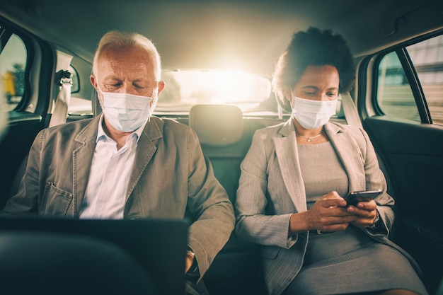 Ujęcie dwóch odnoszących sukcesy wieloetnicznych biznesmenów w masce ochronnej, korzystających z laptopa i smartfona, siedzących na tylnym siedzeniu samochodu podczas porannych dojazdów do pracy podczas pandemii COVID-19.