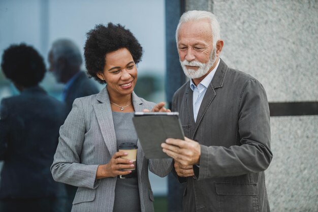 Ujęcie dwóch odnoszących sukcesy wieloetnicznych ludzi biznesu, którzy używają cyfrowego tabletu i prowadzą dyskusję podczas przerwy na kawę przed biurowcem.