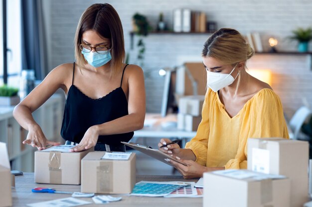 Ujęcie dwóch pięknych niezależnych kobiet biznesu noszących higieniczną maskę na twarz podczas sprawdzania kolejności produktów podczas pakowania i zamykania kartonów w swojej małej firmie rozpoczynającej działalność.