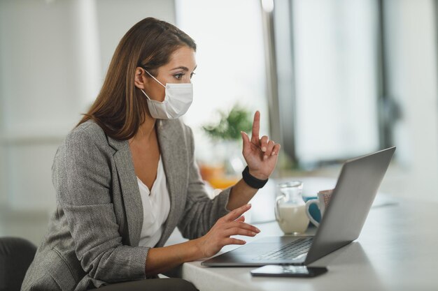 Ujęcie młodej kobiety biznesu w masce ochronnej, siedzącej samotnie w swoim domowym biurze i pracującej na laptopie podczas pandemii COVID-19.