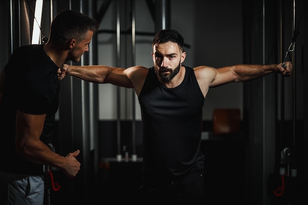 Ujęcie muskularnego faceta w stroju sportowym ćwiczącego z osobistym trenerem na siłowni. Wykonuje ciężkie ćwiczenia na klatkę piersiową na maszynie linowej podczas treningu siłowego.