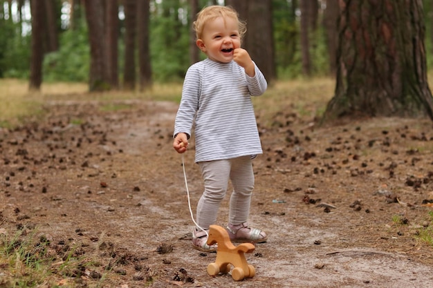 Ujęcie na zewnątrz szczęśliwej dziewczynki w pasiastej koszuli z blond kręconymi włosami bawiącej się w lesie w wiosenny lub letni dzień, trzymającej śmiejącą się drewnianą zabawkę w kształcie konia