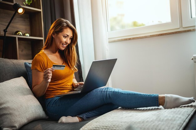 Ujęcie słodkiej młodej kobiety robi zakupy online na swoim laptopie, siedząc na kanapie w domu.