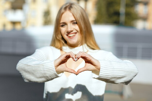 Ujęcie stylu życia: szczęśliwa blondynka w białym swetrze z dzianiny robi symbol serca rękami heart