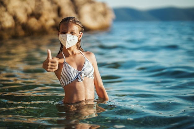 Ujęcie szczęśliwego nastolatka noszącego maskę chirurgiczną, cieszącego się wakacjami na plaży i pokazującego kciuk w górę podczas COVID-19. Patrząc na aparat.