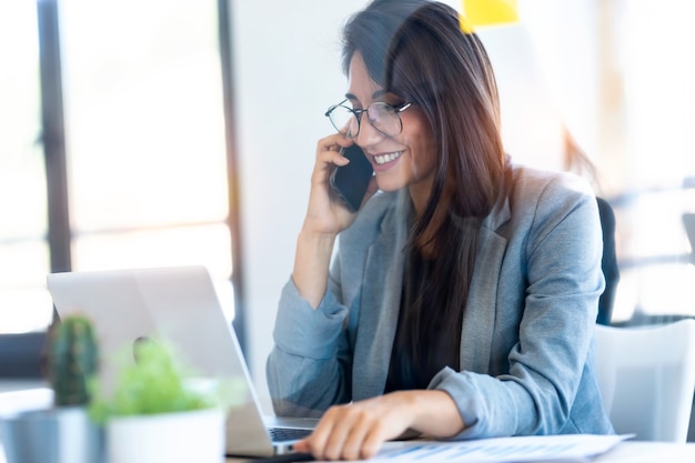 Ujęcie Uśmiechnięta Businesswoman Rozmawia Przez Telefon Komórkowy Podczas Pracy Z Laptopem W Biurze.