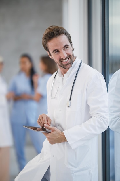 Ujęcie uśmiechniętego lekarza korzystającego z cyfrowego tabletu i zamyślonego patrzącego przez okno podczas szybkiej przerwy w szpitalnym korytarzu.