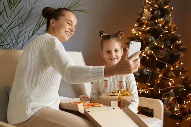 Ujęcie uśmiechniętej kaukaskiej kobiety w białym swetrze siedzącej na kaszlu w pobliżu choinki z uroczą córką jedzącą fast foody, cieszące się pizzą i robiące selfie przez telefon komórkowy