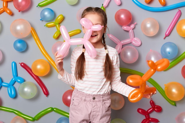 Ujęcie w pomieszczeniu małej dziewczynki z warkoczami ubranej w zwykłe ubrania pozowanie na białym tle na szarym tle z balonami trzymającymi balony z powietrzem postacie zwierząt grające na imprezie
