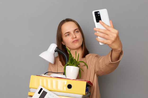 Ujęcie w pomieszczeniu śmiesznej kobiety ubranej w stylową beżową kurtkę trzymającej rzeczy biurowe pozującej się na szarym tle robiącej selfie po zwolnieniu lub rozmowie wideo opowiadającej o zwolnieniu