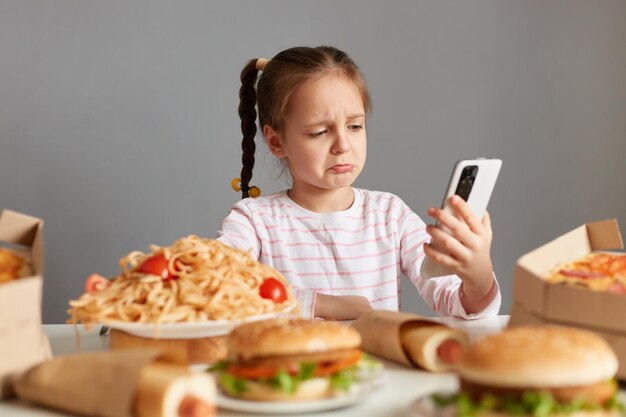 Ujęcie w pomieszczeniu smutnej płaczącej dziewczynki z warkoczykami siedzącej przy stole pizza hot dogi hamburgery rozmawiające przez połączenie wideo utrzymuje dietę nie może jeść pysznych fast foodów odizolowanych na szarym tle