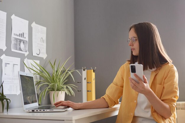 Ujęcie w pomieszczeniu uroczej brązowowłosej kaukaskiej kobiety freelancer siedzącej przy biurku pracującej na komputerze i używającej telefonu komórkowego patrzącej na ekran w żółtej koszuli praca online