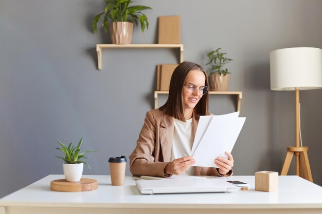 Ujęcie w pomieszczeniu uroczej, optymistycznej kobiety o brązowych włosach, ubranej w beżową kurtkę, pracującej w biurze z dokumentami wyrażającymi pozytywne emocje, sprawdzającymi rachunki