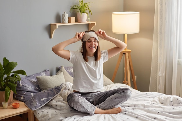 Ujęcie w pomieszczeniu uśmiechniętej, radosnej kobiety noszącej maskę do spania rano, siedzącej na łóżku i zdejmującej opaskę, cieszącej się doskonałym nastrojem