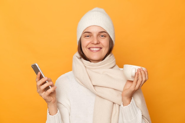 Ujęcie w pomieszczeniu uśmiechniętej, szczęśliwej, zadowolonej kaukaskiej kobiety w ciepłej czapce i szaliku, pozującej się na białym tle na żółtym tle, trzymającej kubek kawy za pomocą smartfona