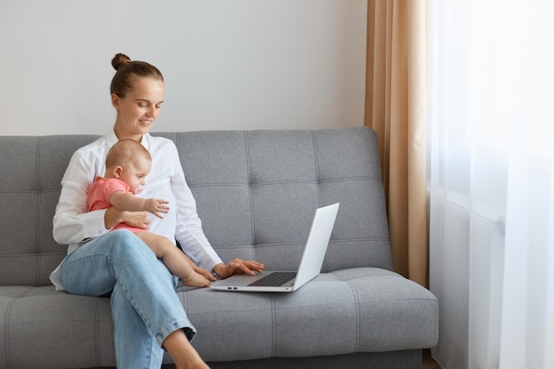 Ujęcie w pomieszczeniu uśmiechniętej zadowolonej kobiety w białej koszuli i dżinsach siedzącej na kanapie z córeczką i podającej jej wodę z butelki, pracującej na laptopie