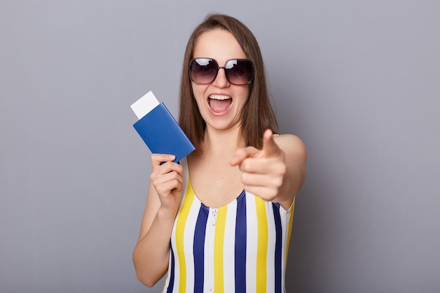 Ujęcie w pomieszczeniu uszczęśliwionej kobiety w stroju kąpielowym i okularach przeciwsłonecznych, trzymającej paszport i wskazującej na aparat uśmiechający się szeroko, zapraszającej na wycieczkę reklamową biura podróży na białym tle na szarym tle