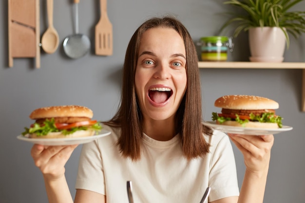 Ujęcie w pomieszczeniu zdumionej uśmiechniętej kobiety trzymającej dwa pyszne świeże hamburgery z soczystym pieczonym kurczakiem i sałatką z rukoli, która jest bardzo głodna i krzyczy wow ze szczęścia, ciesząc się niezdrowym jedzeniem