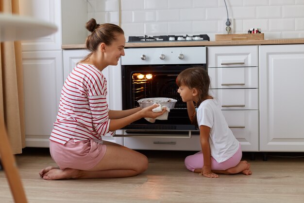 Ujęcie Z Profilu W Pomieszczeniu Uśmiechnięta Biorąca Pieczenie Z Piekarnika, Jej Córka Stojąca W Pobliżu I Pachnąca Pysznymi Słodyczami, Dziecko Pomagające Matce W Kuchni, Rodzina Pozująca W Domu Siedząc Na Podłodze.