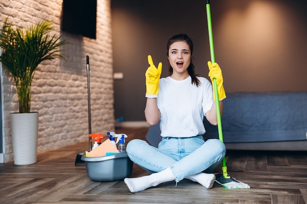 Ujęcie zdziwiona sprzątaczka w żółtej rękawicy trzyma mopa w salonie