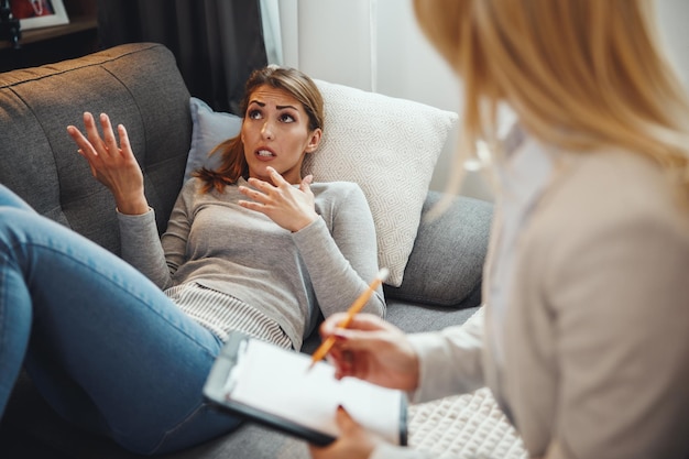 Ujęcie zestresowanej młodej kobiety rozmawiającej ze swoją psychoterapeutką siedzącą na sofie w salonie.