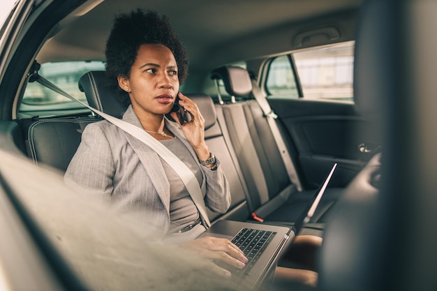 Ujęcie zmartwionej czarnej bizneswoman rozmawiającej przez smartfona, siedząc na tylnym siedzeniu samochodu podczas porannej jazdy do pracy.